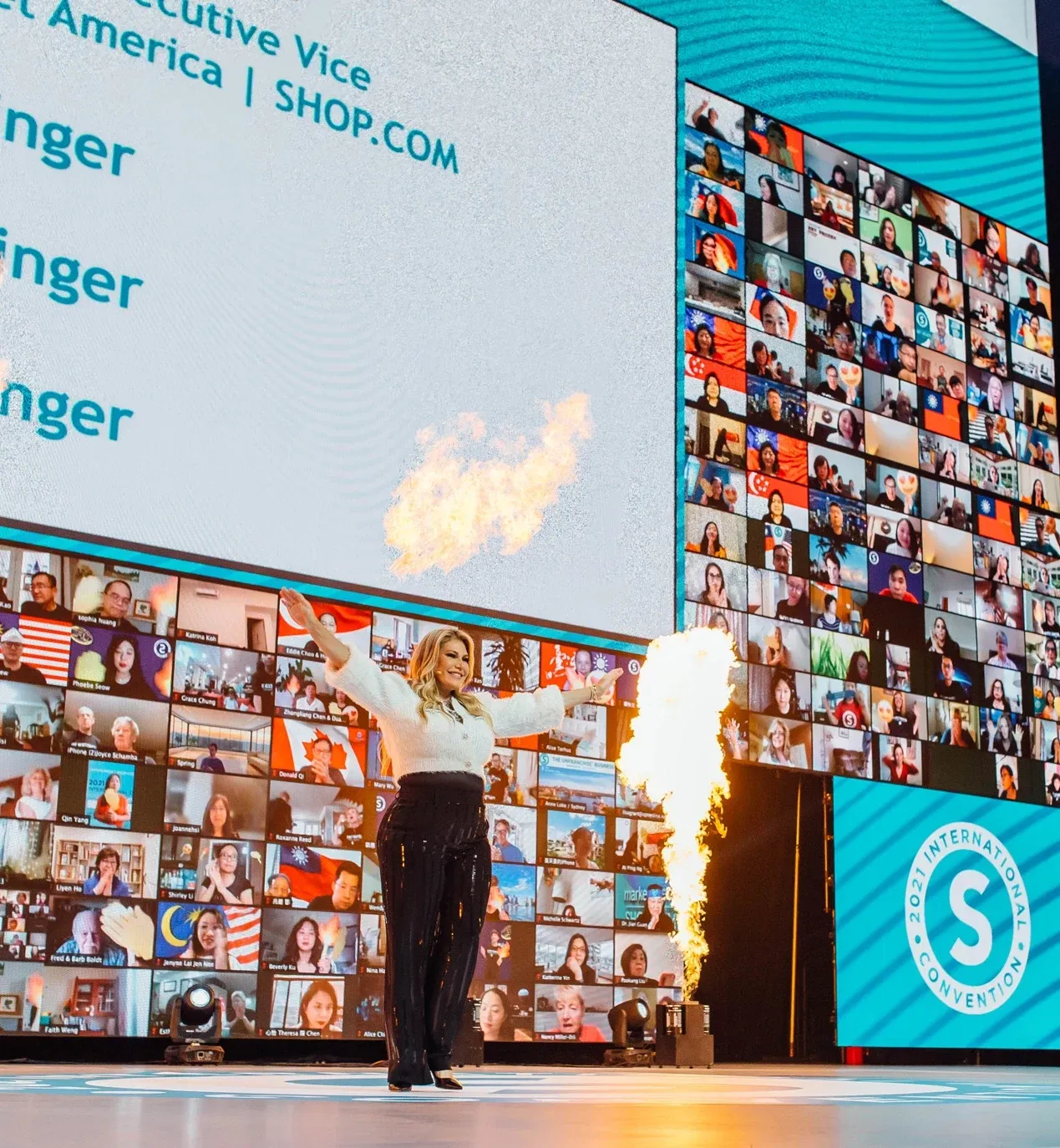 Speaker spreads her arms as a flame jet fires beside an LED wall of virtual attendees
