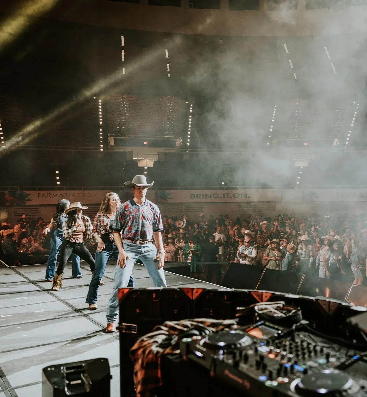 Dancers in cowboy hats line-dance on stage beside a DJ booth as a western-dressed crowd watches