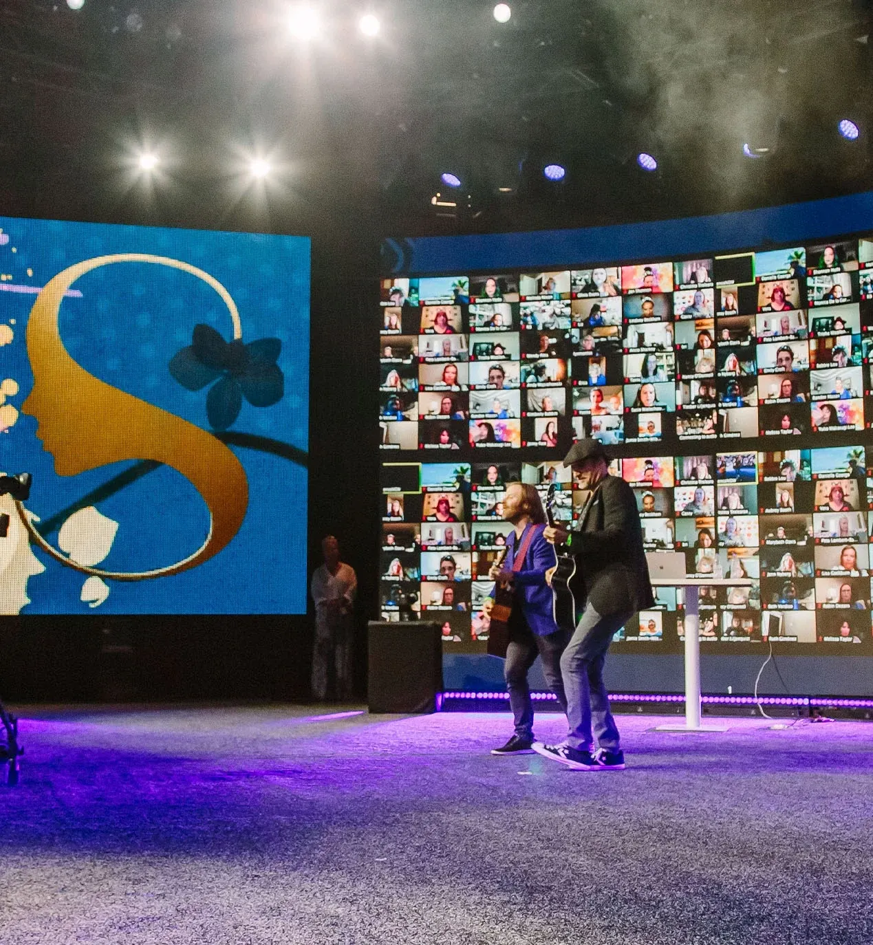 Two guitarists perform on a purple-lit set beside a SeneGence logo screen and a wall of virtual attendees