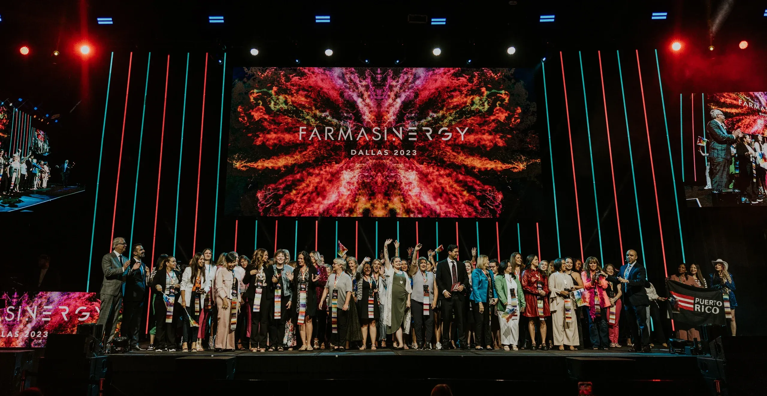 Large group of honorees celebrates on stage beneath a FARMASINERGY Dallas 2023 screen and LED strips