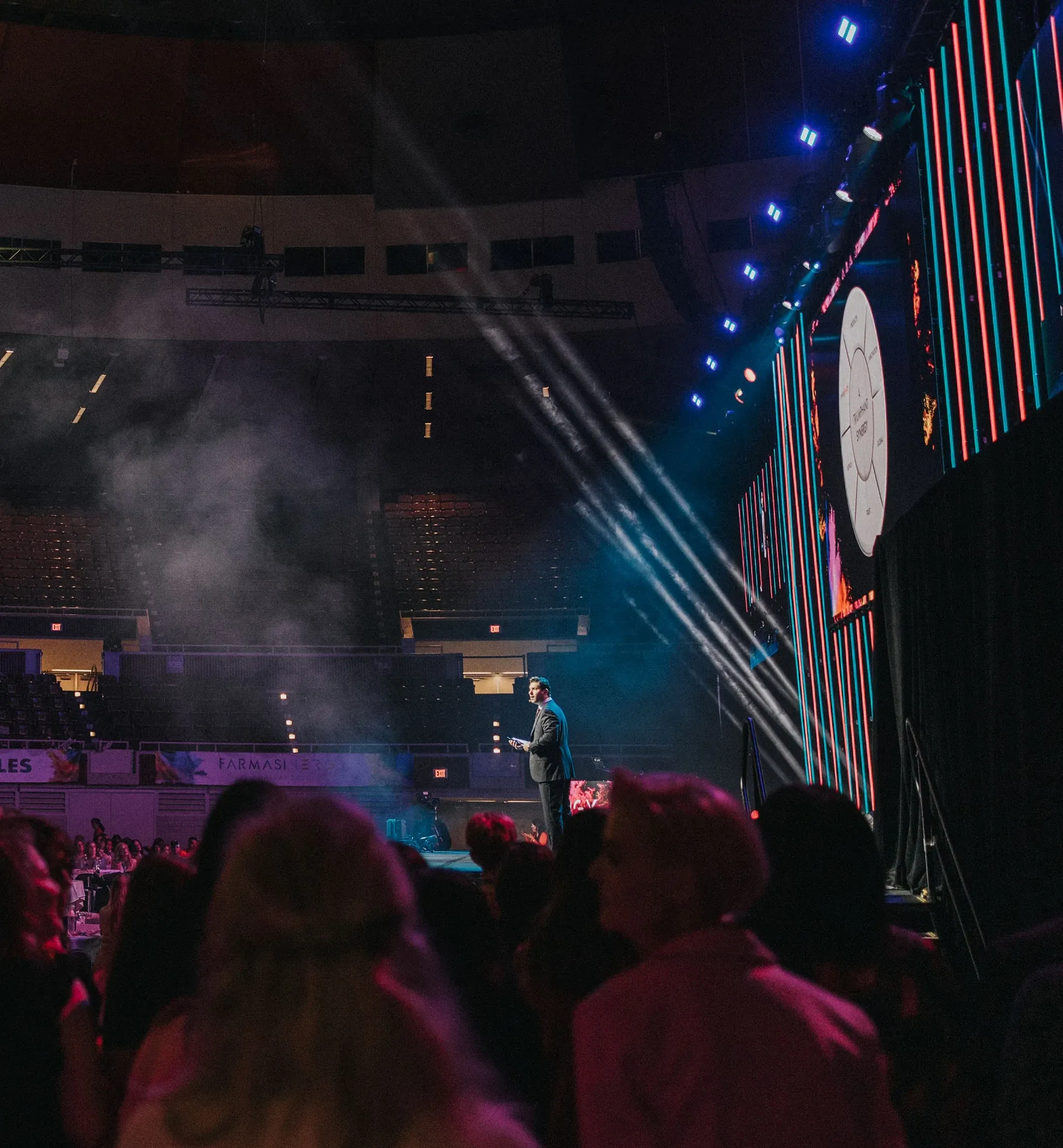 Suited speaker addresses the arena floor beside towering red and teal LED columns and a slide screen