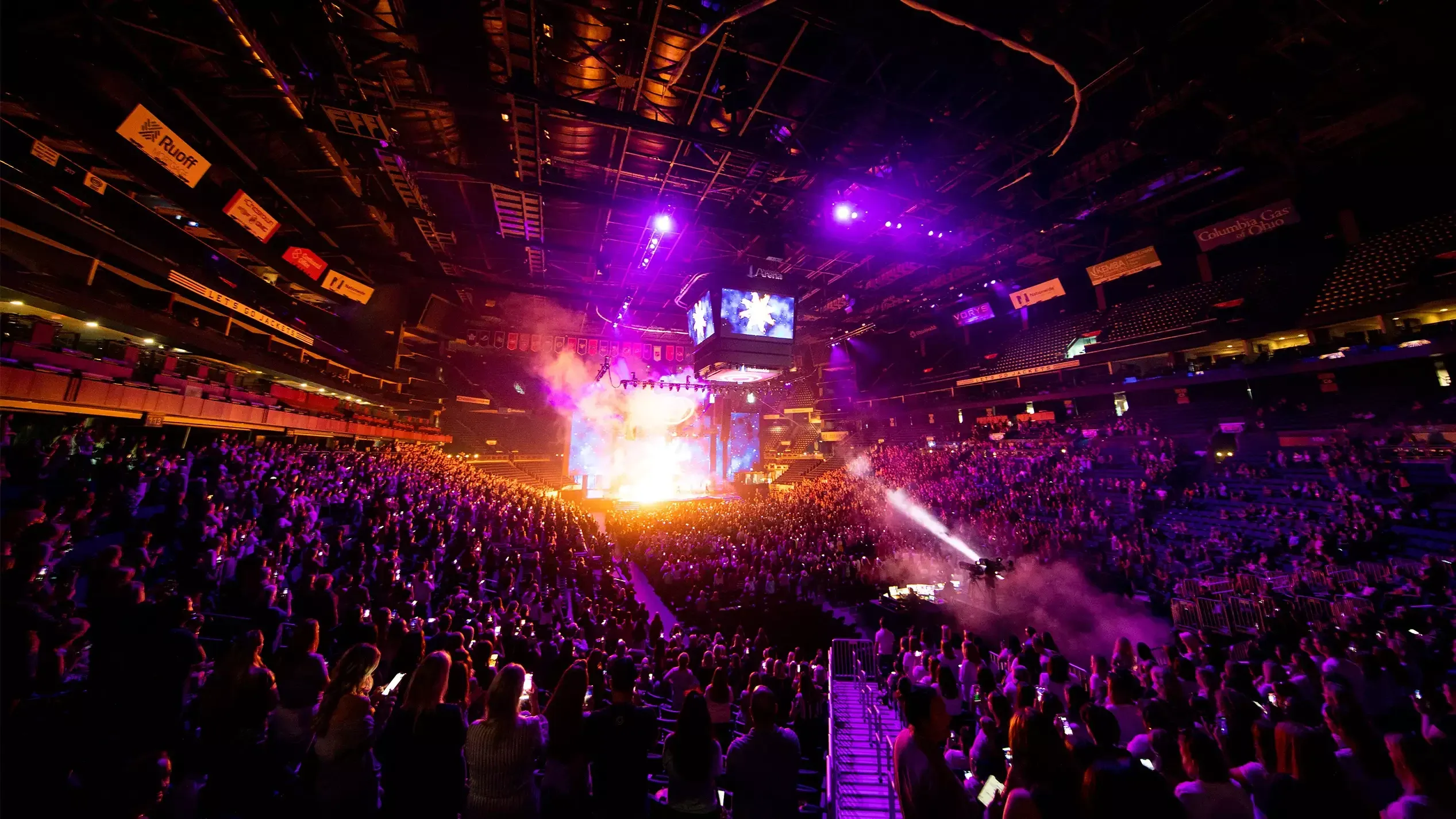 Packed arena holding up phone lights as pyro and haze erupt from a stage wrapped in video screens