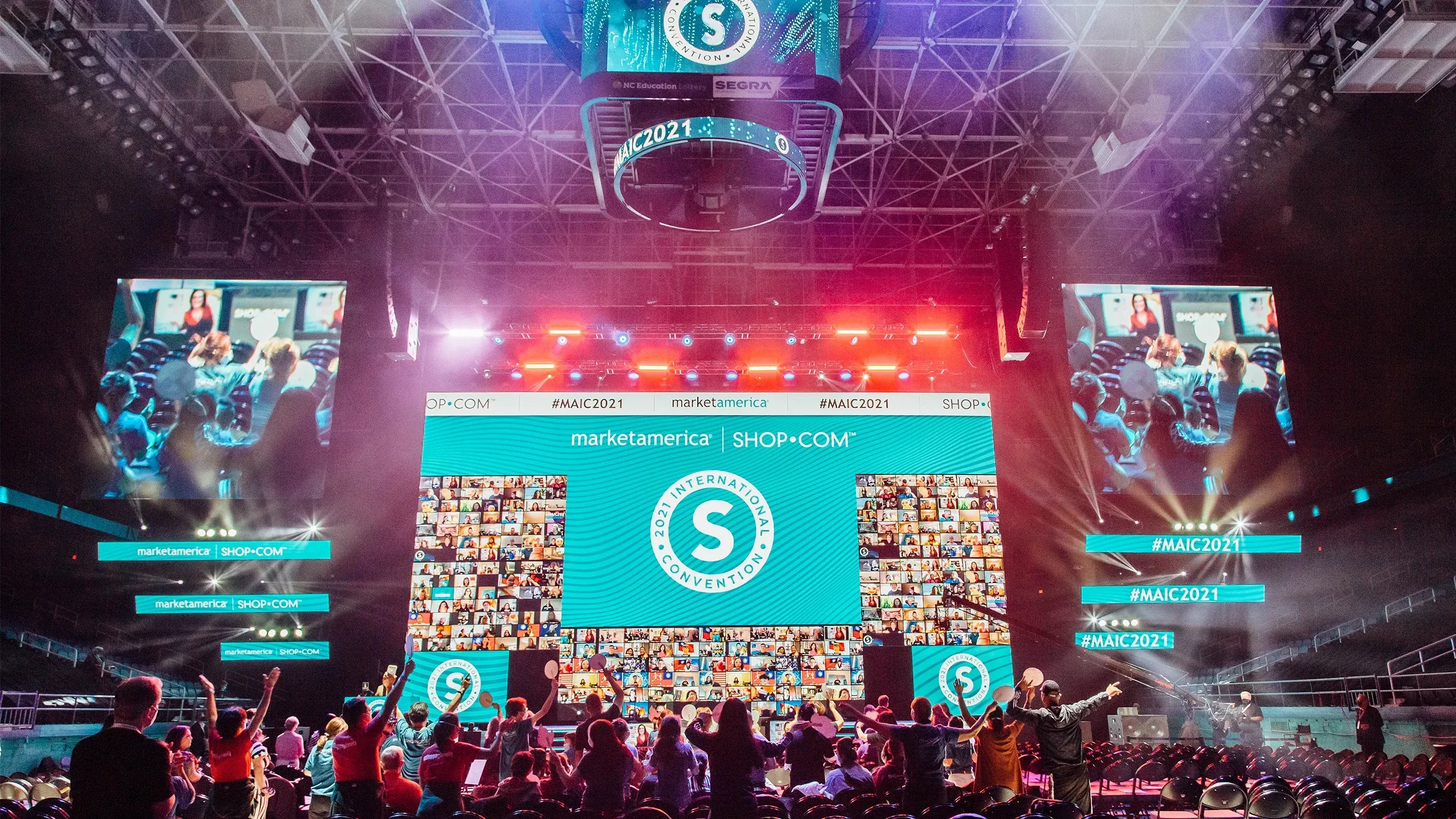 In-person attendees cheer beneath Market America 2021 International Convention screens mixing logos and virtual attendees