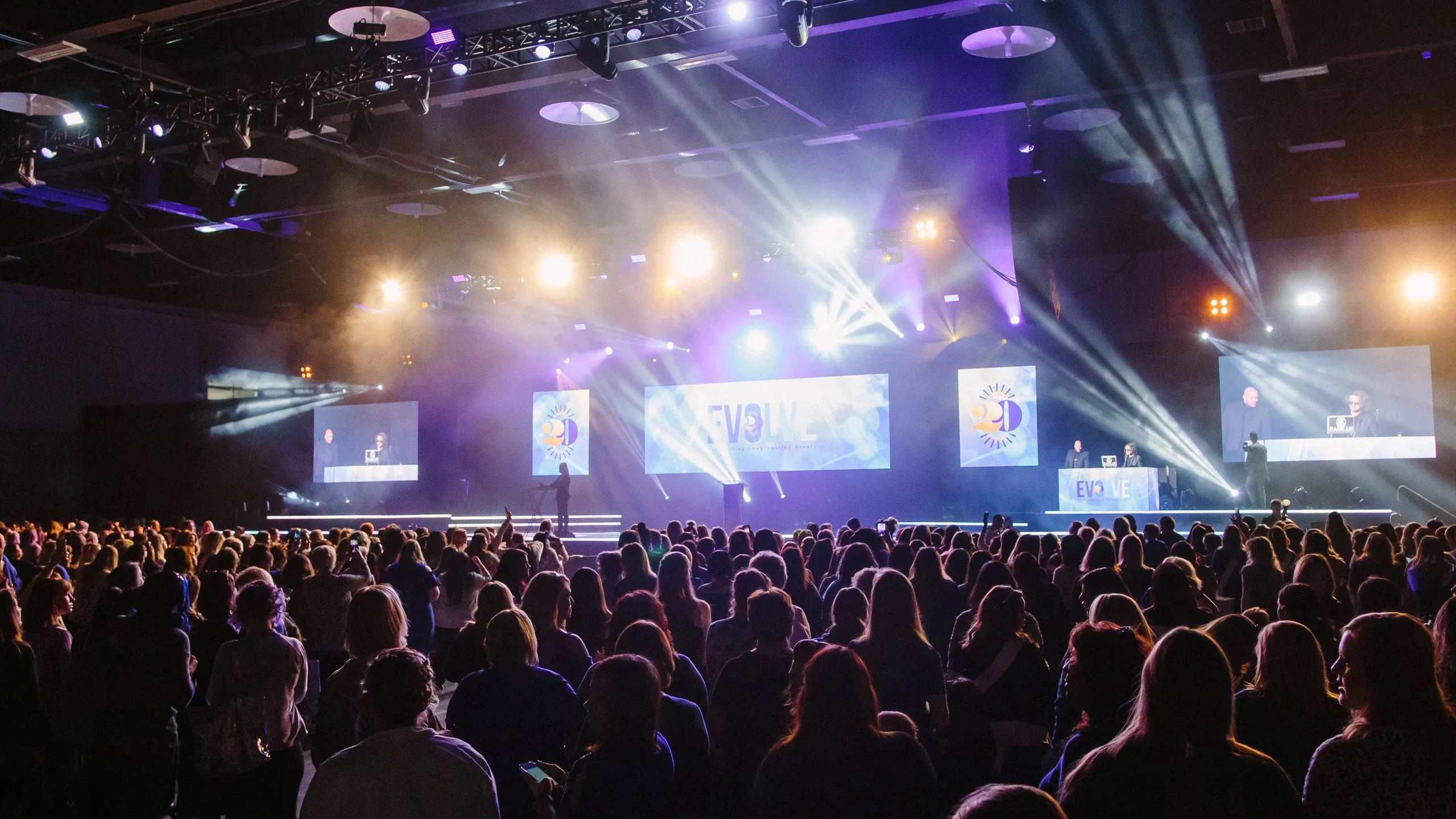 Crowd fills a convention hall facing a stage with EVOLVE and 20 graphics, beam lighting and IMAG screens