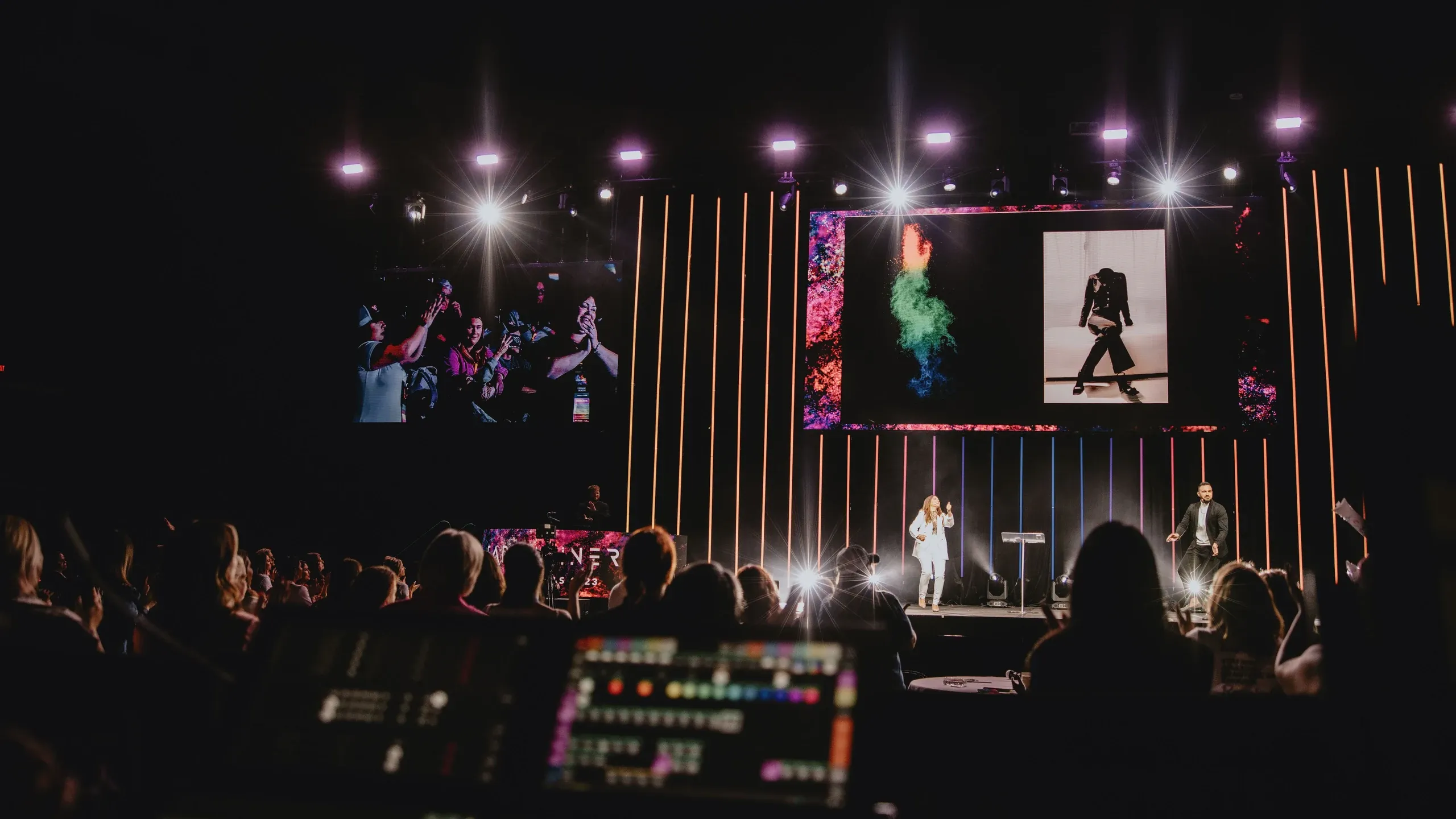 View from the lighting console of two presenters on stage below video screens and starburst lights