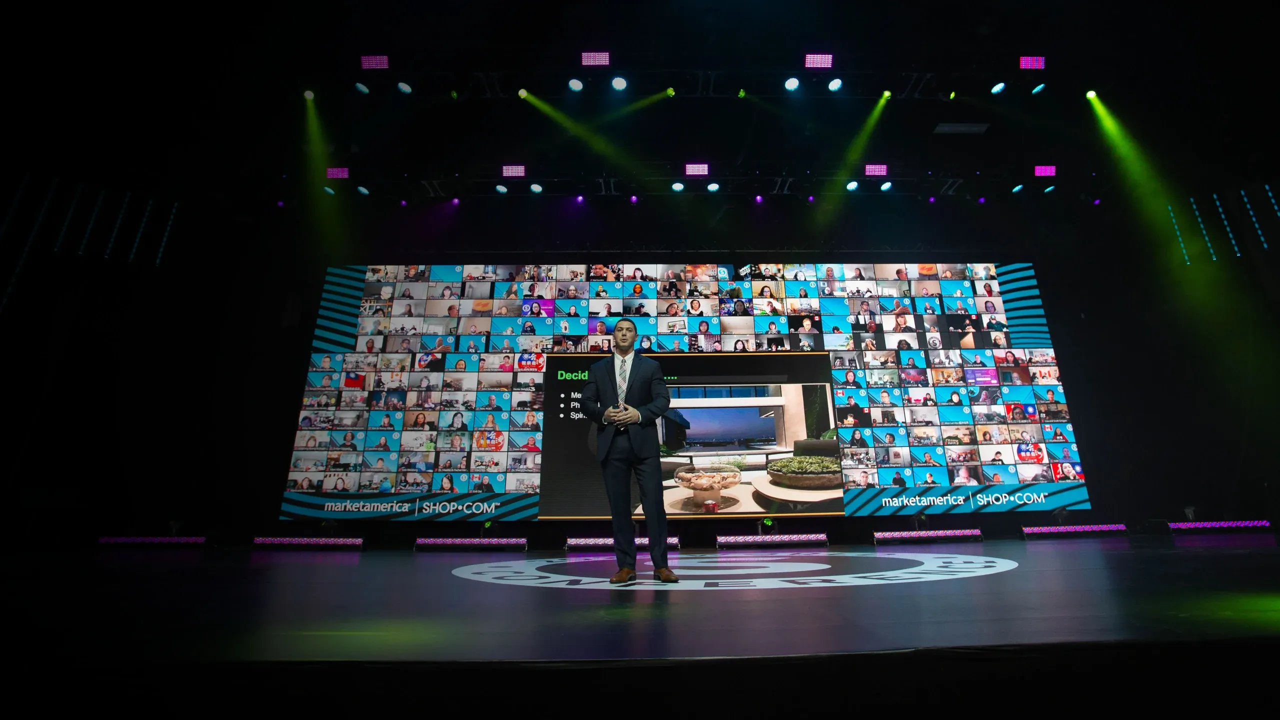 Suited speaker stands center stage before a wide LED wall of remote attendees and a presentation slide